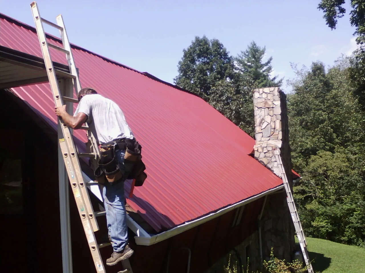 Roofer on a ladder beside a red metal roof and stone chimney — Soffit & Fascia Repair by Roofing Brothers in Red Bud